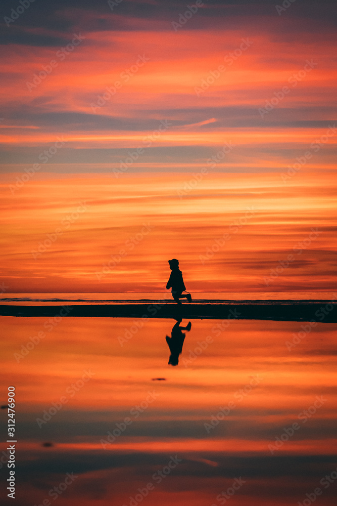Little girl in a hat running on the sea shore in Baltics with a red and orange sky at the sunset