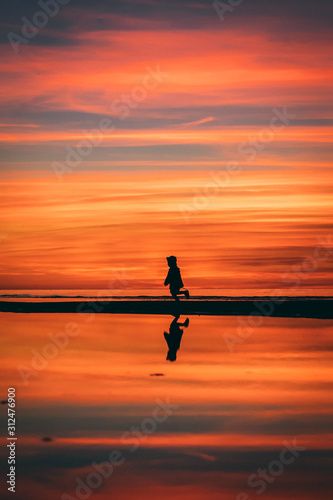 Little girl in a hat running on the sea shore in Baltics with a red and orange sky at the sunset © Sergejs