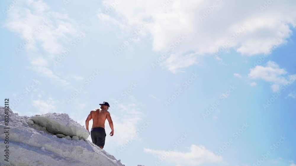 Shirtless man going down the hill. Back view of muscular sportsman in the mountains under blue sky. View from below. Slow motion. Shirtless man going down the hill. Back view of muscular sportsman in the mountains under blue sky. View from below. Slow motion.