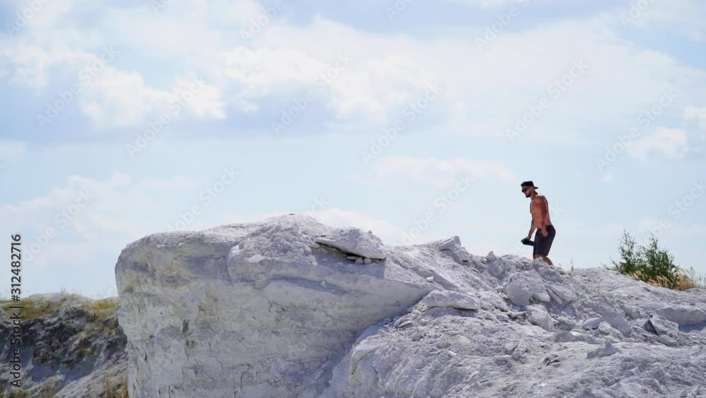 Slow motion of strong athlete in the mountains. Shirtless sportsman walking up the white hill in summer. Muscular man exercises outdoors.