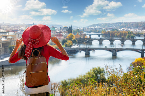 Fotografie A traveler woman with red sunhat sits on a hill above Prague, Czech Republic, an