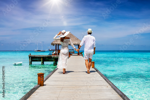 Canvas-taulu A happy couple in white summer clothing on vacation walks along a wooden pier ov