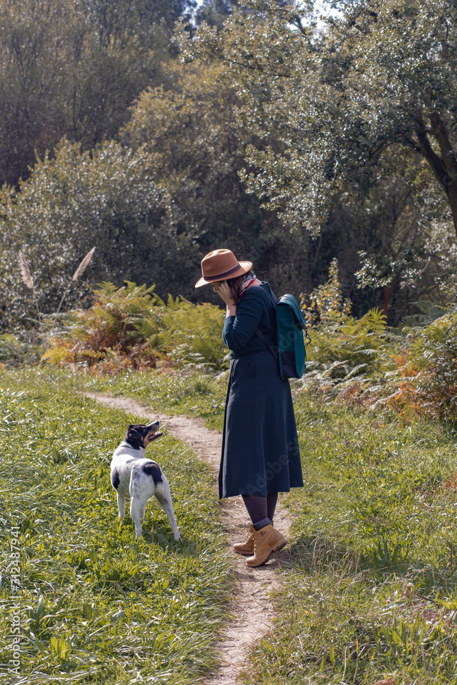 young woman with hat standing next to her dog in colorful forest 