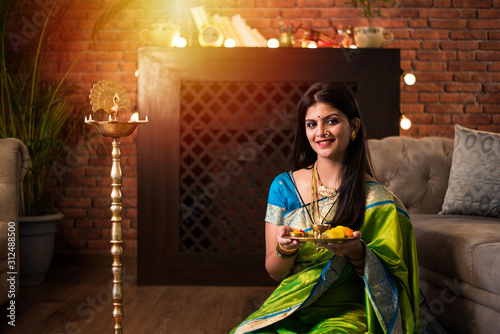 Indian Girl / women holding Pooja Thali while wearing green saree and sitting at home with flower rangoli and Samai, celebrating Diwali or hindu festival. selective focus