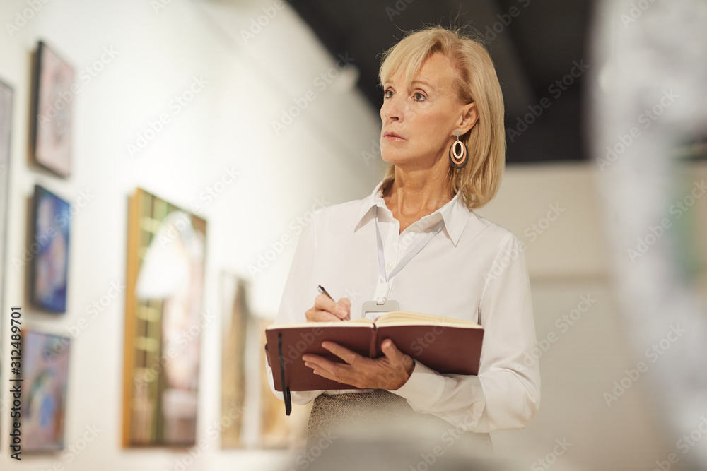 Waist up portrait of female art expert appraising paintings in gallery and taking notes, copy space Waist up portrait of female art expert appraising paintings in gallery and taking notes, copy space