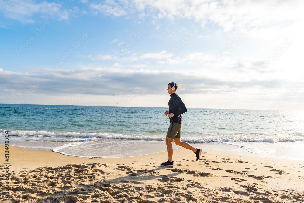 Fit male runner training on the summer beach and listen to music against beautidul sky and sea Fit male runner training on the summer beach and listen to music against beautidul sky and sea