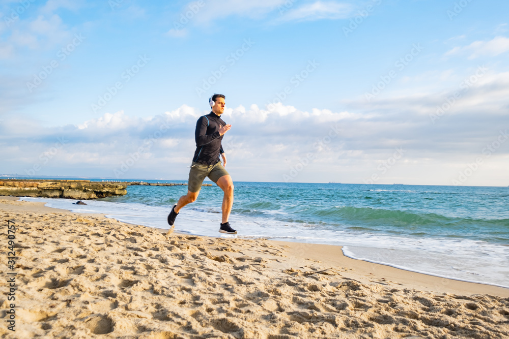 Fit male runner training on the summer beach and listen to music against beautidul sky and sea	