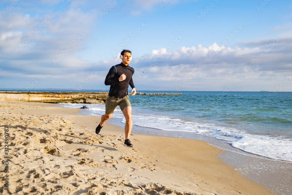 Fit male runner training on the summer beach and listen to music against beautidul sky and sea Fit male runner training on the summer beach and listen to music against beautidul sky and sea