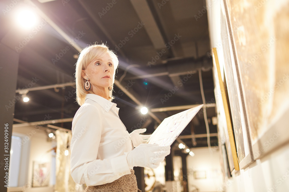 Low angle portrait of female art expert holding painting while appraising works in modern gallery or museum, copy space Low angle portrait of female art expert holding painting while appraising works in modern gallery or museum, copy space