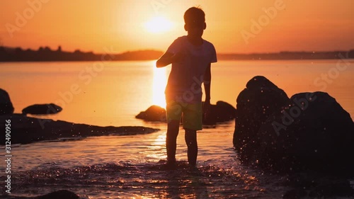 Dressed boy in water at sunset. Silhouette of a happy child making splashes of water in slow motion near stones. Beautiful orange sunset over the river.