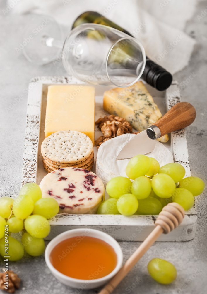 Glass and bottle of white wine with selection of various cheese in wooden box and grapes on wooden table background. Blue Stilton, Red Leicester and Brie Cheese and Cheddar with nuts and honey. Glass and bottle of white wine with selection of various cheese in wooden box and grapes on wooden table background. Blue Stilton, Red Leicester and Brie Cheese and Cheddar with nuts and honey.