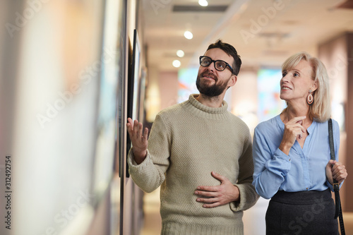 Waist up portrait of smiling mature couple looking at paintings while enjoying exhibition in art gallery or museum, copy space