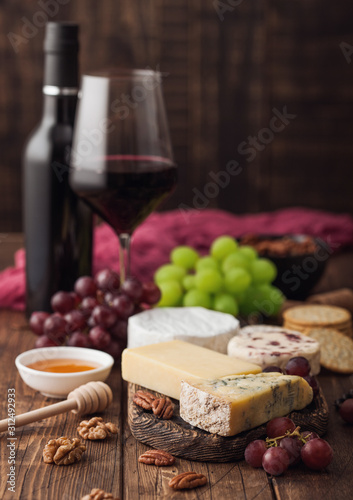 Glass and bottle of red wine with selection of various cheese on the board and grapes on wooden background. Blue Stilton, Red Leicester and Brie Cheese and bowl of nuts and crackers.