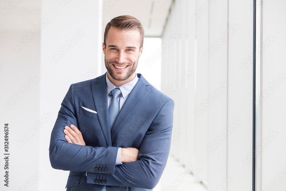 Portrait of smiling handsome young businessman standing with arms crossed in new office - obrazy, fototapety, plakaty 