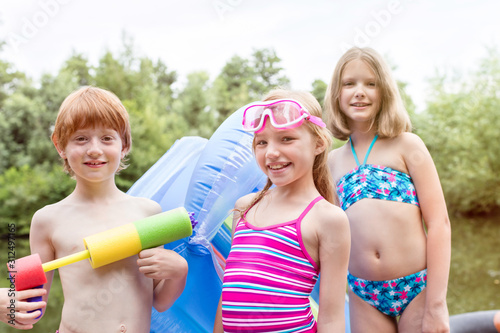 Photo Portrait of smiling friends in swimwear standing with pool raft and squirt gun a