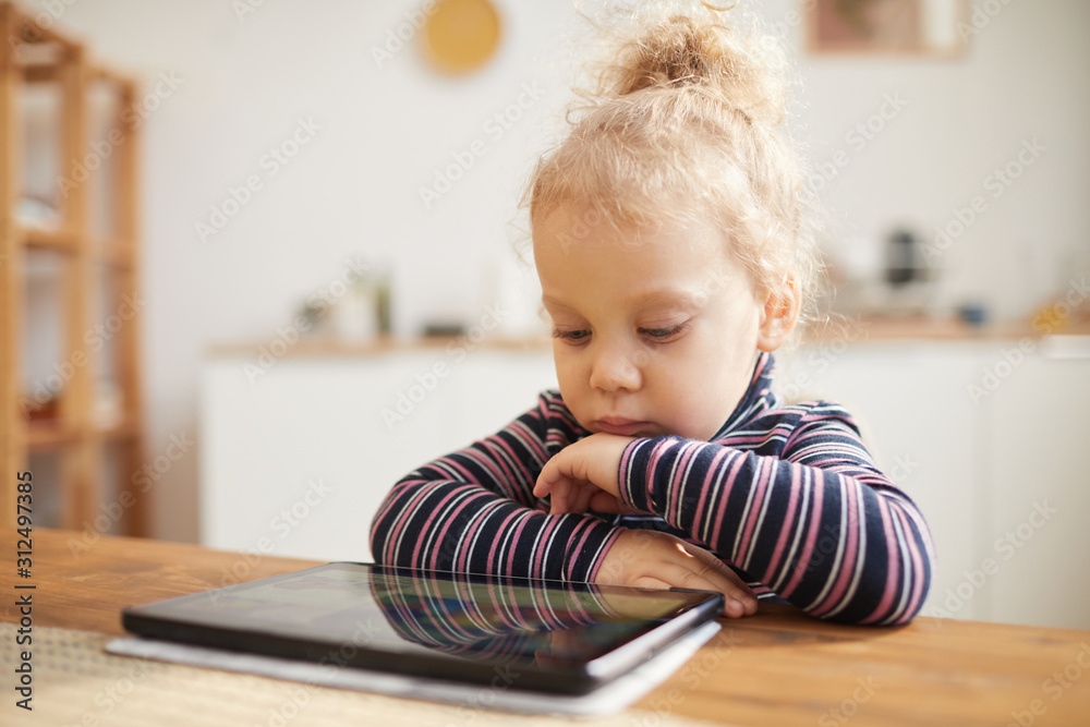 Warm-toned portrait of cute little girl looking at digital tablet while sitting at big table in cozy kitchen, copy space Warm-toned portrait of cute little girl looking at digital tablet while sitting at big table in cozy kitchen, copy space