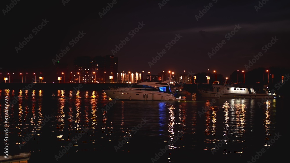Luxurious modern private yacht and boat at the pier at night. Ship crossing skyline in the harbor in summer awesome nightly. Colorful lights of city glow shine reflected in water wave at nighttime.