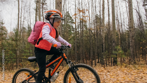 One caucasian children rides bike road in autumn park. Little girl riding black orange cycle in forest. Kid goes do bicycle sports. Biker motion ride with backpack and helmet. Mountain bike hardtail. © ivandanru