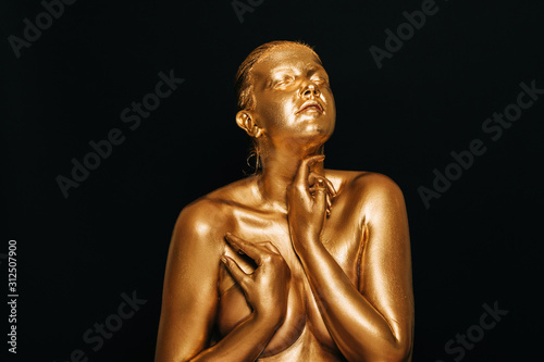 Portrait of sensual woman covered with golden paint, posing on black background, eyes closed