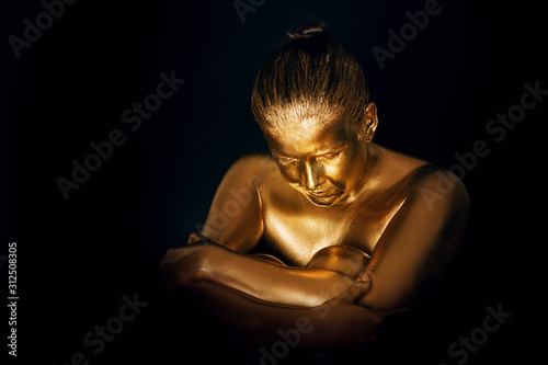 Portrait of sensual woman covered with golden paint, posing on black background, eyes closed, head down