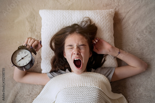 Sleepy teenager girl turning alarm off in the morning while lying in bed 