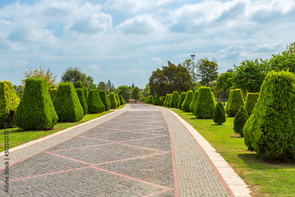 Cobbled walkway in a park with bright green clipped trees, blue sky and beautiful clouds. Regular Park in Adana, Turkey, Merkez Park