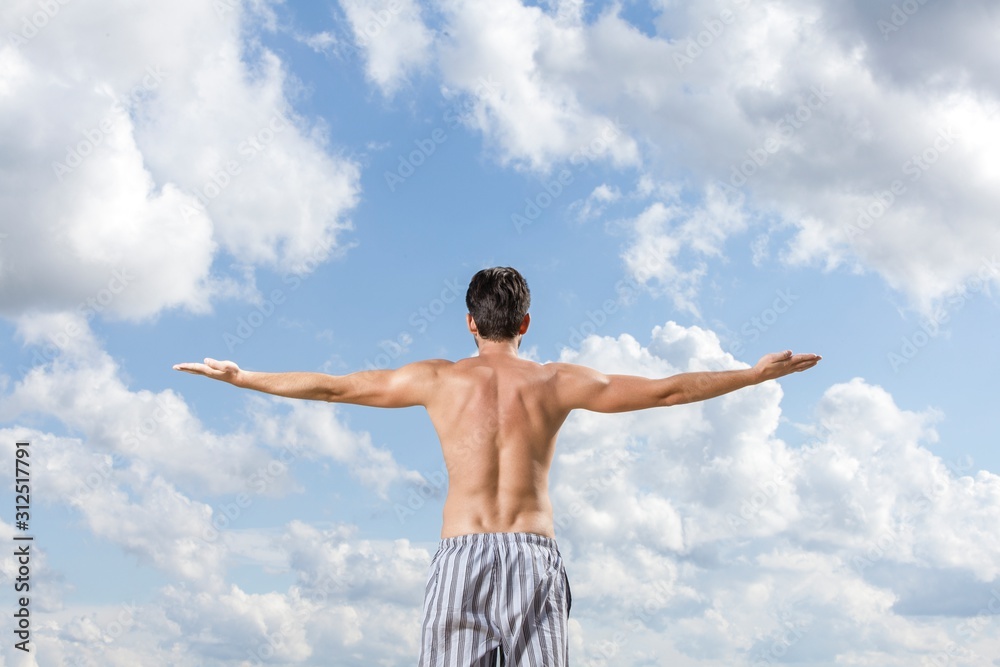 Rear view of shirtless young man standing arms outstretched against cloudy sky Rear view of shirtless young man standing arms outstretched against cloudy sky