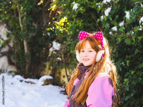 Portrait of little redhead girl with snow and trees in the background. Enjoying winter holidays