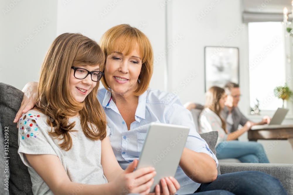 Loving mother and daughter using tablet PC with family sitting in background at home Loving mother and daughter using tablet PC with family sitting in background at home