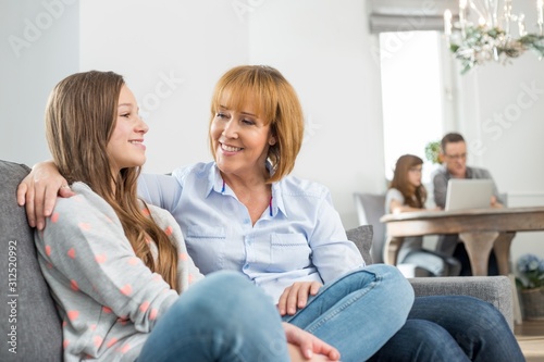 affectionate mother and daughter sitting on sofa with family in background