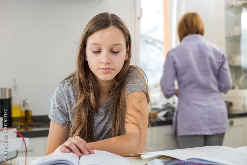 Girl studying at table with mother standing in background Girl studying at table with mother standing in background