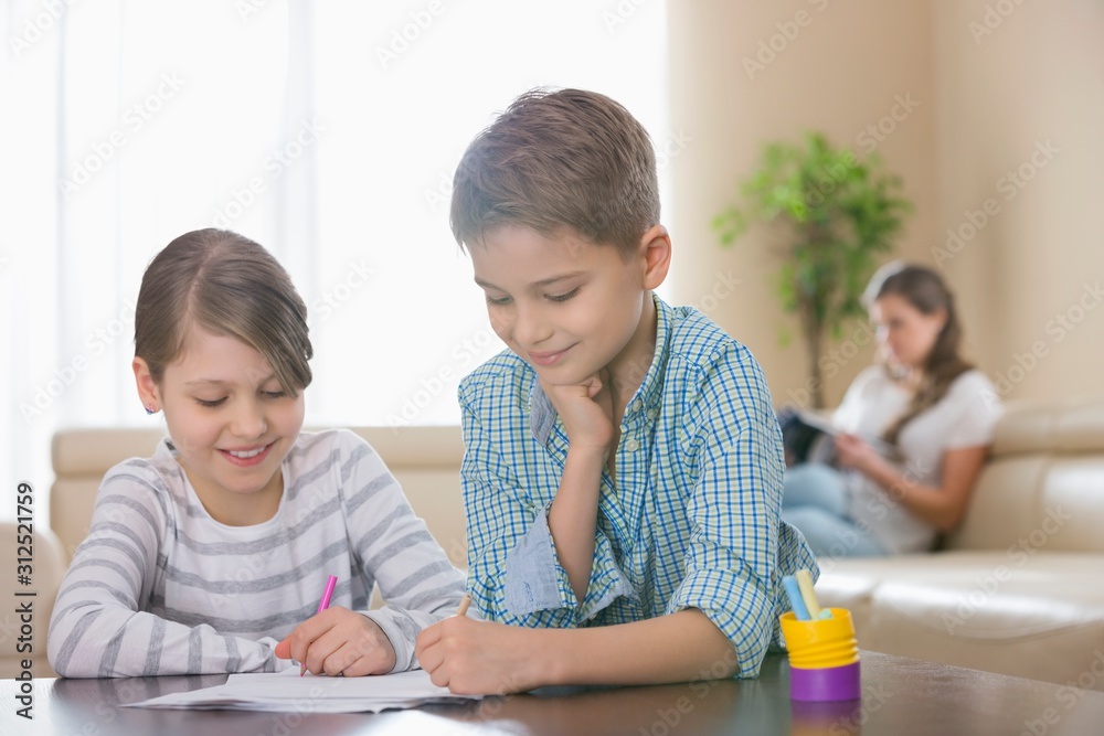 Siblings drawing together at table with mother in background Siblings drawing together at table with mother in background