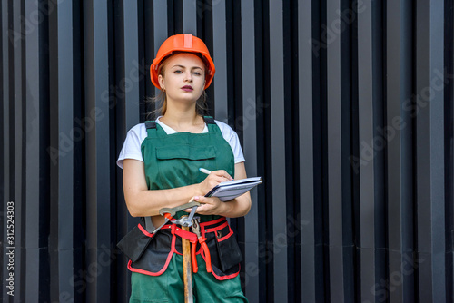 Woman worker in coverall with tool belt posing near grey wall