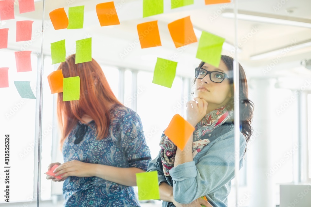 Creative businesswomen reading sticky notes on glass wall in office Creative businesswomen reading sticky notes on glass wall in office