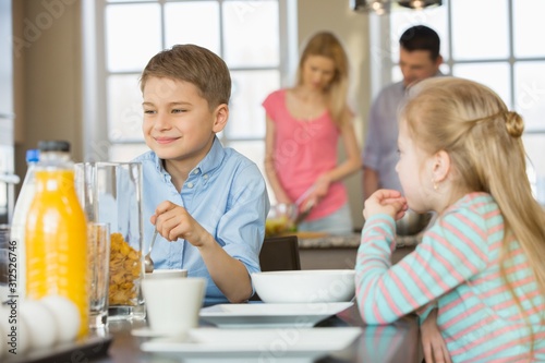 Siblings having breakfast at table with parents cooking in background