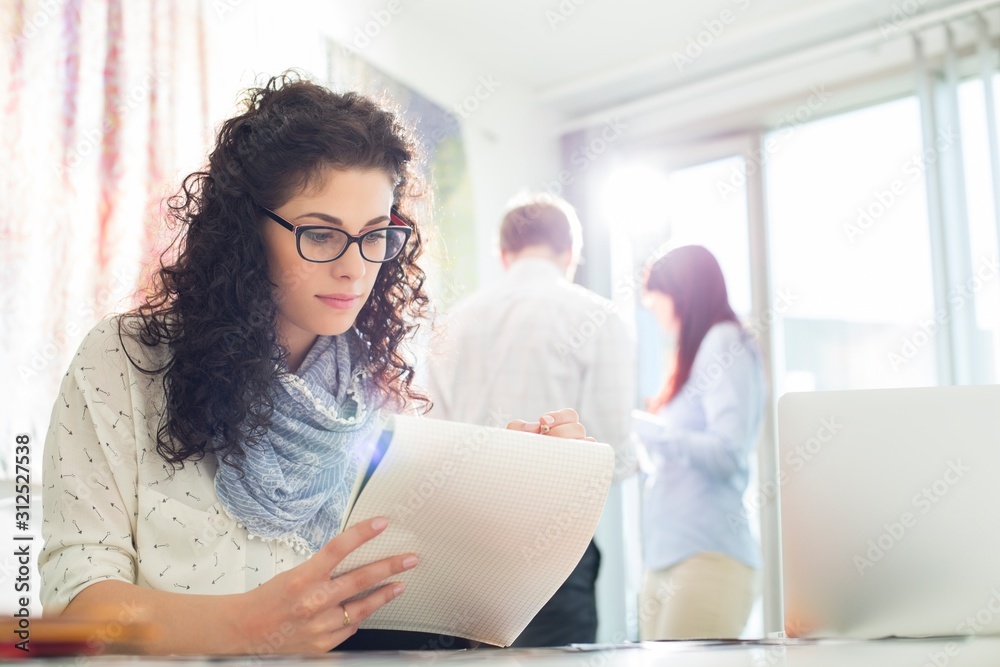 Businesswoman reading notepad at creative office with colleagues in background Businesswoman reading notepad at creative office with colleagues in background