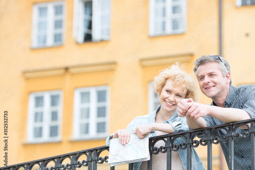 Happy middle-aged couple with map leaning on railing against building