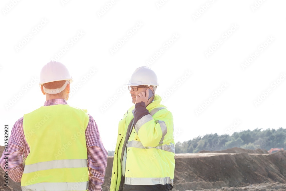 Male engineer using mobile phone while standing with colleague at construction site against clear sky Male engineer using mobile phone while standing with colleague at construction site against clear sky