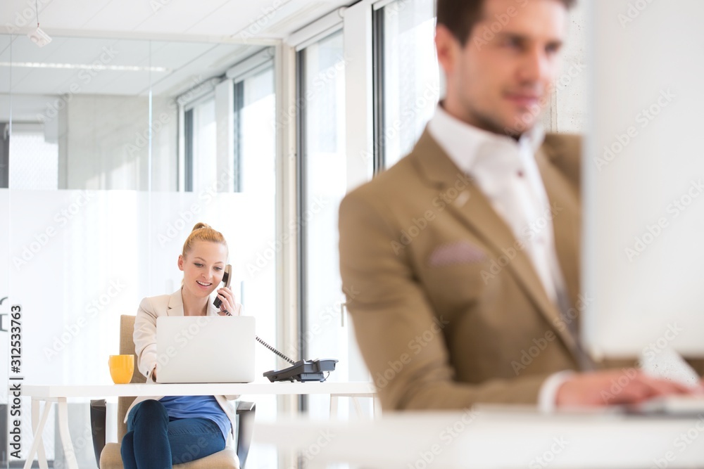Young businessman using landline phone with male colleague in foreground at office Young businessman using landline phone with male colleague in foreground at office