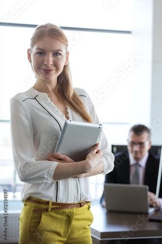 Portrait of confident young businesswoman holding tablet PC with businessman in background at office