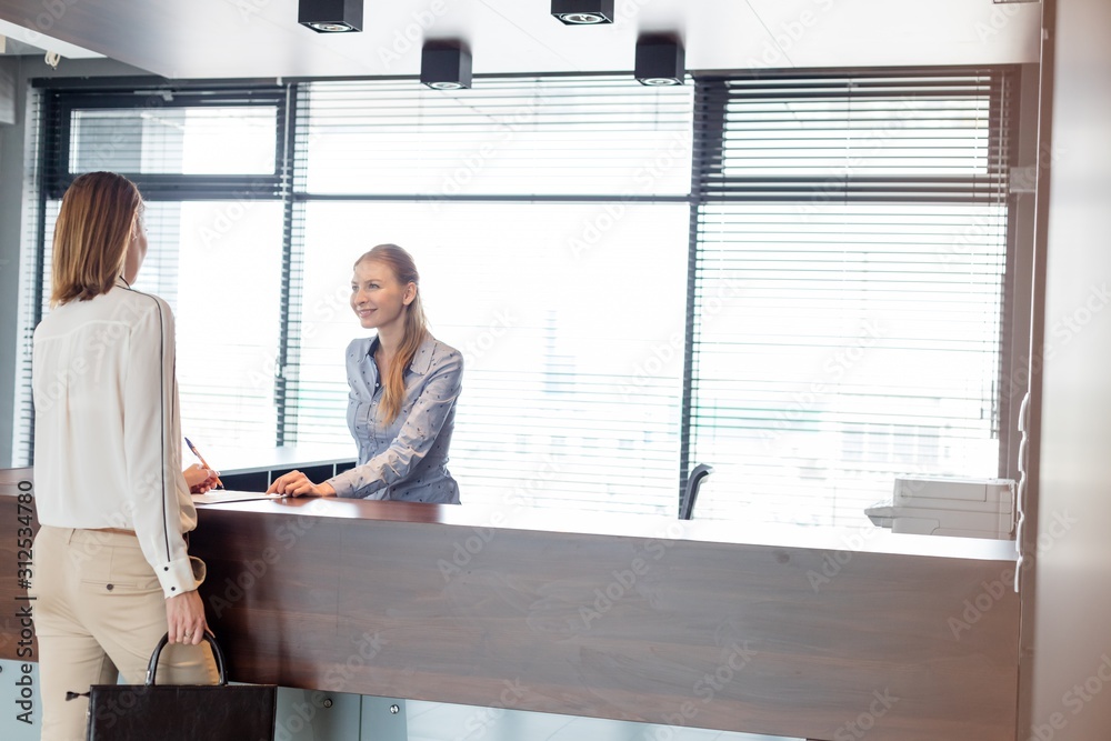 Young female receptionist looking at businesswoman signing document in office
