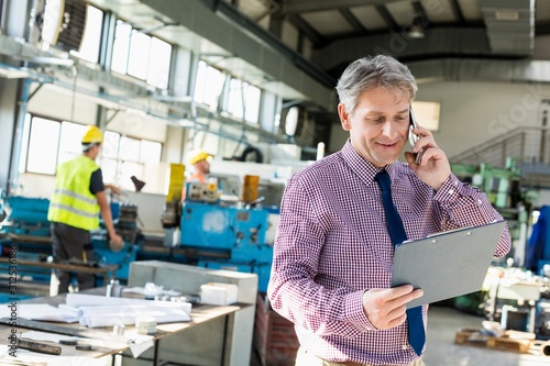 Fotografie Mature male supervisor looking at clipboard while talking on mobile phone in ind