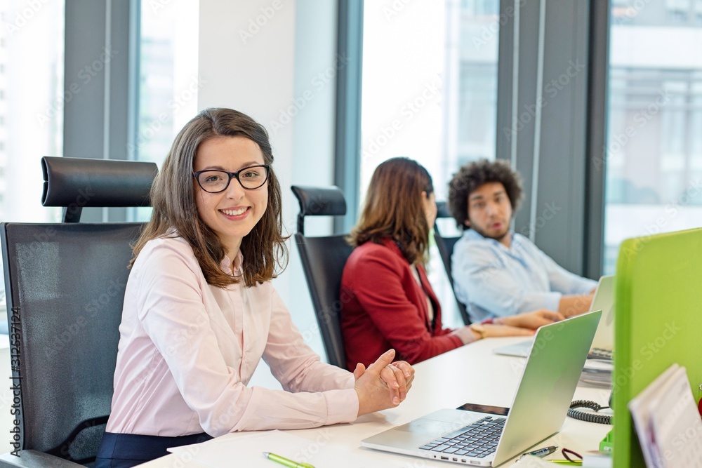 Portrait of smiling young businesswoman with laptop while colleagues in background at office Portrait of smiling young businesswoman with laptop while colleagues in background at office