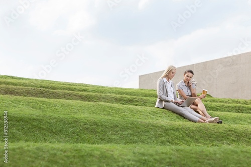 Full length of female business executives with disposable coffee cup and laptop sitting on grass steps against sky