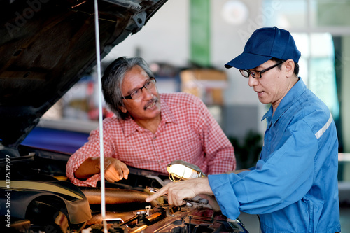 Asian automotive mechanic with blue uniform point to the car engine and explain about the problem to the customer in the garage
