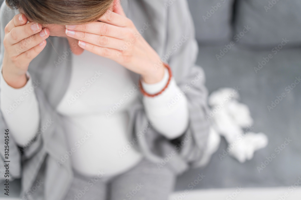 Pregnant woman catching cold, flu, virus, sitting at home on a couch, blowing nose and being sick.