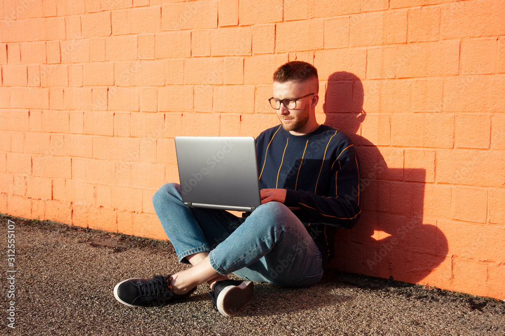 Young caucasian man in casual-wear sitting cross-legged on ground and working on laptop. Freelancer working outside typing on laptop computer. Red wall on the background.