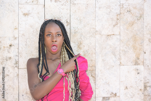 Surprised Afro-style black girl with long black and blondes braids using sunglasses rests on a wall and points to the right with her finger while looks at the camera