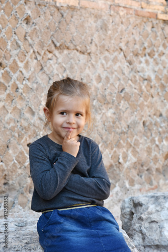 Pensive cunning little girl looking up and thinking about something. Little dreamer in casual wear with finger on lips. Kid having a good idea. Making up a game. Ancient wall in the background. © Davide Zanin