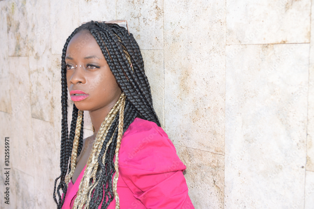 Fototapeta premium Afro-style black girl with long black and blondes braids wearing sunglasses rests on a wall and daydreams